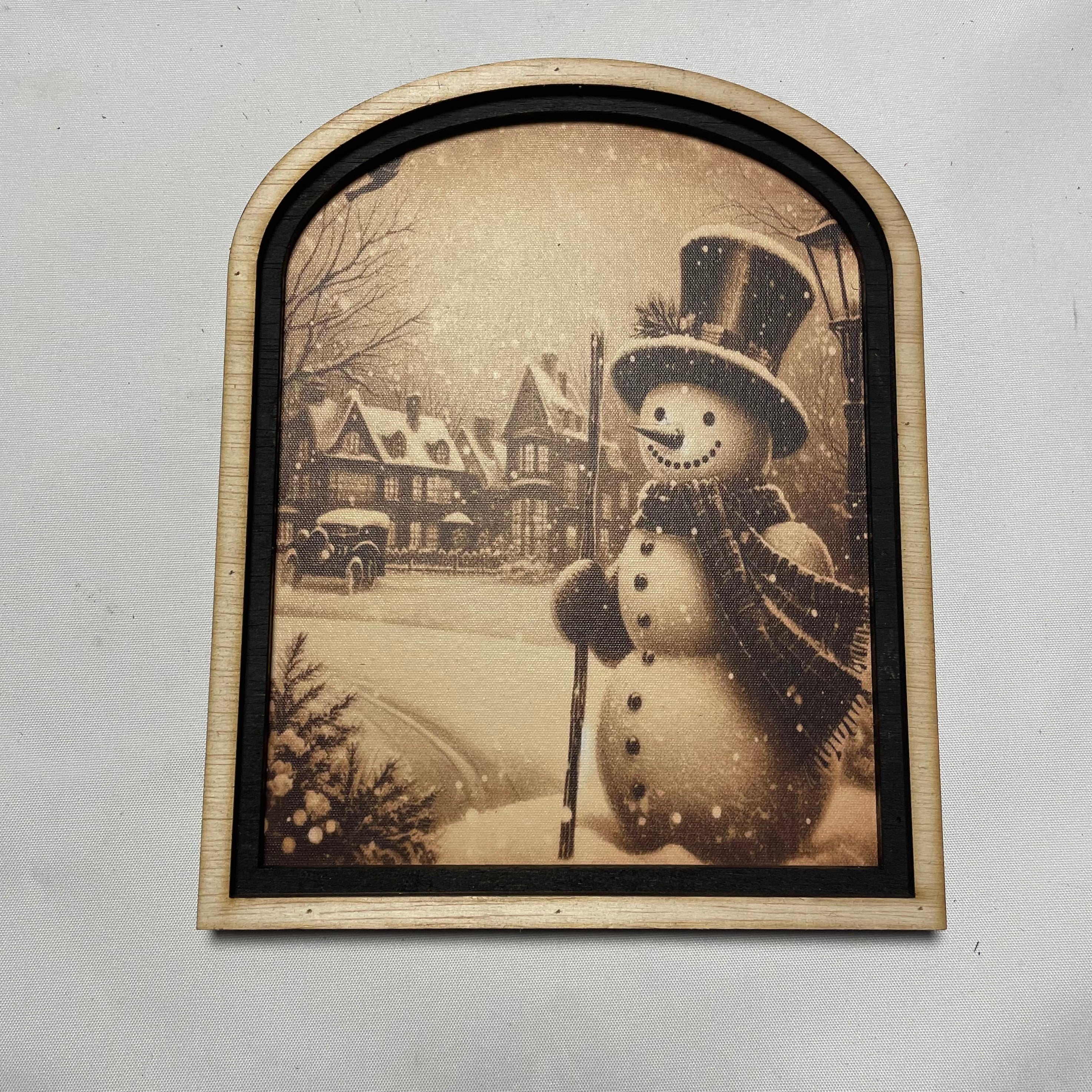 Snowman in snow covered village with two tone arched frame
